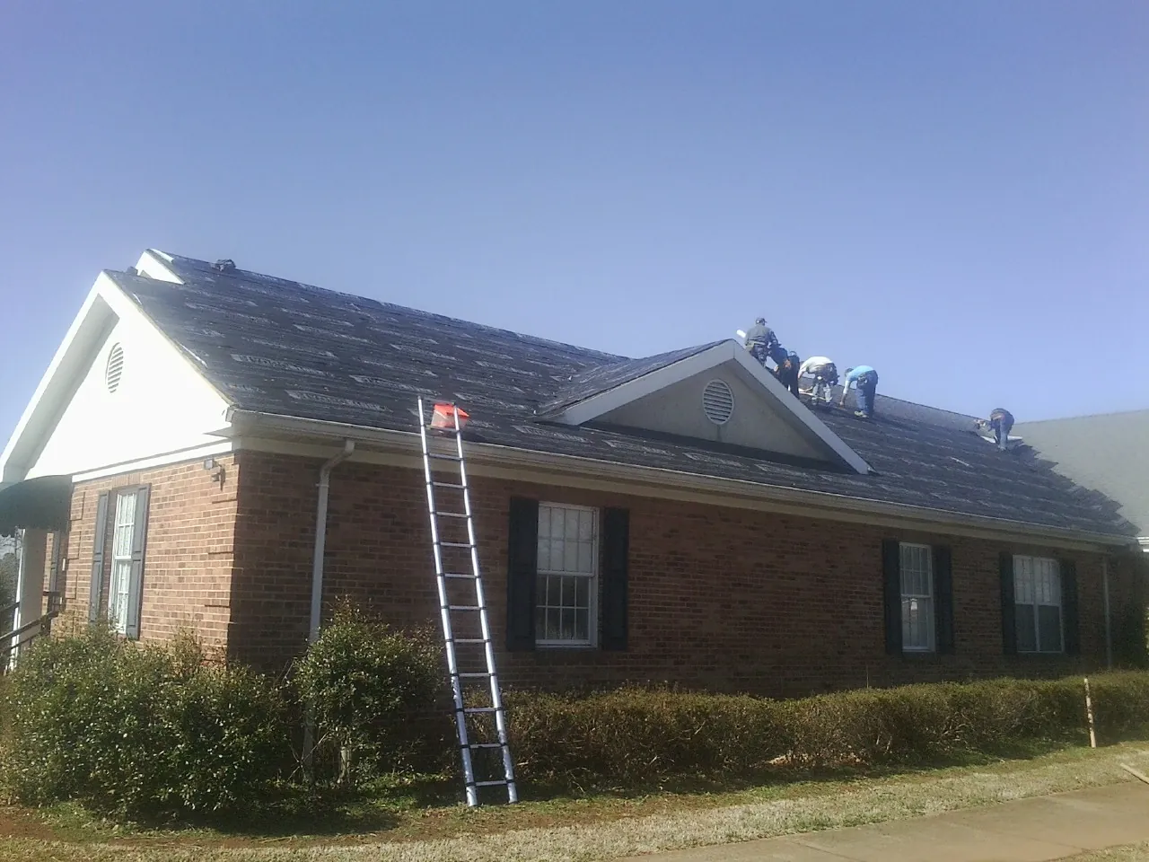 Crew tearing shingles off a brick ranch roof with a ladder set — Metal Roof Repair work by our crew in Indian Hills
