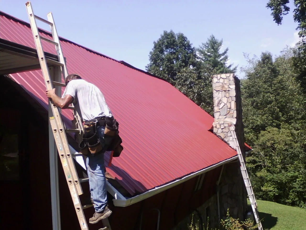 Roofer on a ladder beside a red metal roof and stone chimney — Soffit & Fascia Repair by Roofing Brothers in Indian Hills